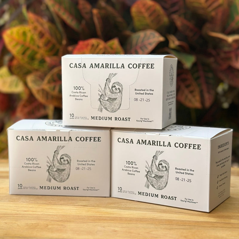 Three boxes of Casa Amarilla Coffee Sloth Blend pods on a wooden surface with tropical foliage in the background